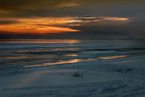 A serene winter landscape at sunset, with icy waters and a colorful sky reflecting on the snow-covered shore.