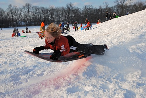 A child sleds down a snowy hill, surrounded by other kids enjoying winter activities.