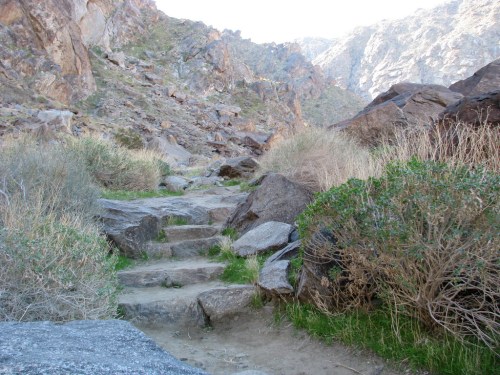 A rocky path winds through a mountainous landscape, surrounded by sparse vegetation and rugged terrain.