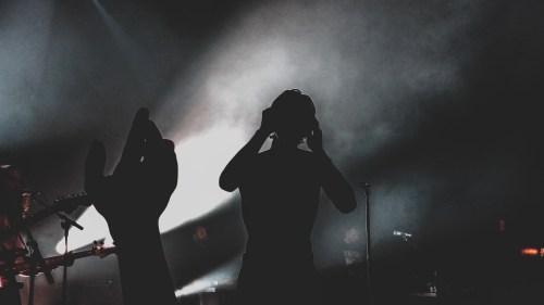 Silhouettes of a person and a hand against a smoky, illuminated stage during a concert.