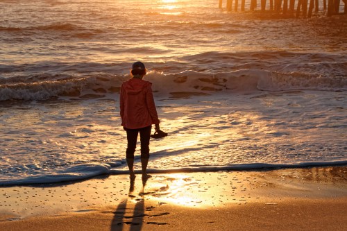 A person stands on a beach at sunset, holding shoes, with waves gently lapping at their feet and a pier in the background.