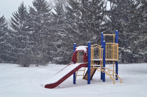 A colorful playground slide partially buried in snow, surrounded by tall trees in a winter landscape.
