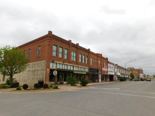 A quiet street lined with historic brick buildings under a cloudy sky, featuring trees and greenery along the sidewalk.