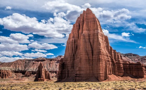 A towering red rock formation under a blue sky with fluffy clouds, surrounded by desert landscape.