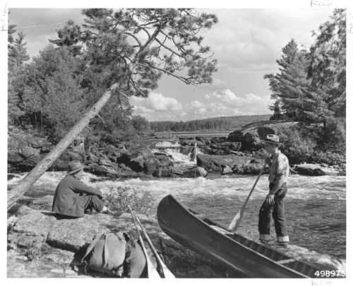 Two men by a river, one sitting on a rock and the other paddling a canoe near a waterfall and trees.