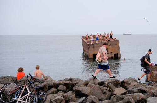 A group of people swim and relax on a concrete structure in the water, while others walk along rocky shorelines.