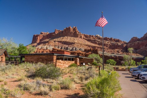 A stone building with an American flag in front, surrounded by desert vegetation and red rock formations in the background.