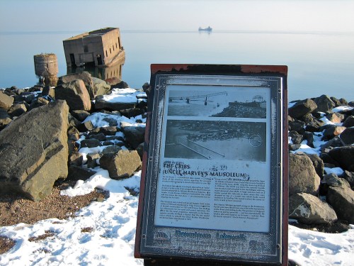 A historical sign near a snowy shoreline, with a sunken structure and a distant ship on calm waters.