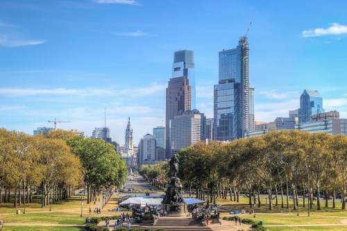 A city skyline featuring modern skyscrapers, green trees, and a clear blue sky in an urban park setting.