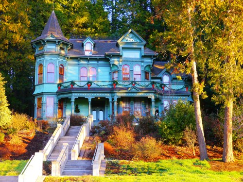 A colorful Victorian-style house adorned with holiday decorations, surrounded by trees and greenery.