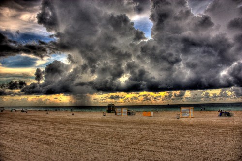 A beach scene with dark clouds overhead, a distant rain shower, and colorful beach huts along the sandy shore.