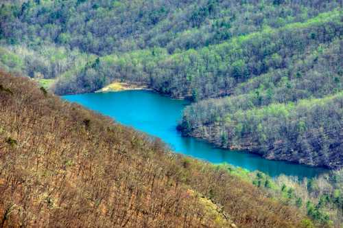 Aerial view of a turquoise lake surrounded by green and brown hills in a mountainous landscape.