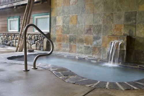 A serene hot tub with a stone wall and a small waterfall, surrounded by a rustic wooden structure.