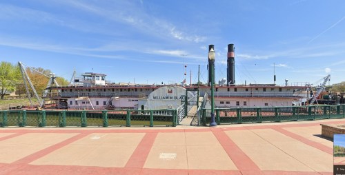 A historic riverboat, the William M. Black, docked by a waterfront with blue skies and greenery in the background.