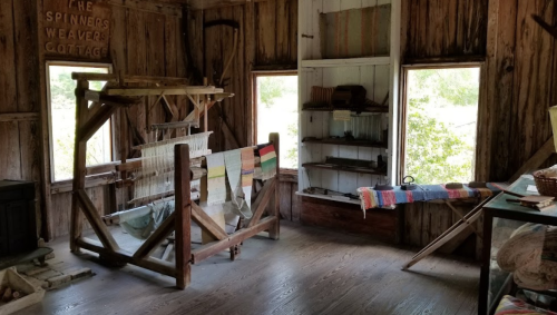 Interior of a rustic cottage with a loom, shelves, and colorful woven textiles, surrounded by wooden walls and windows.