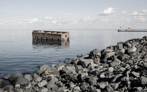 A sunken structure partially submerged in calm water, with a rocky shoreline and a distant lighthouse on the horizon.