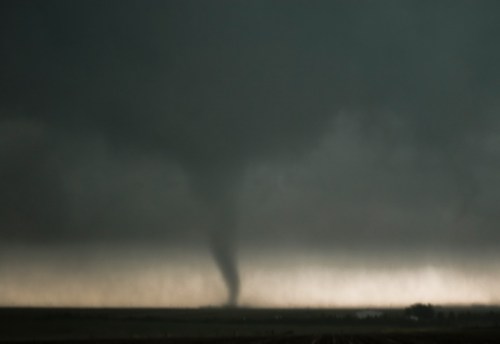 A dark tornado descends from a stormy sky over a flat landscape, with ominous clouds surrounding it.