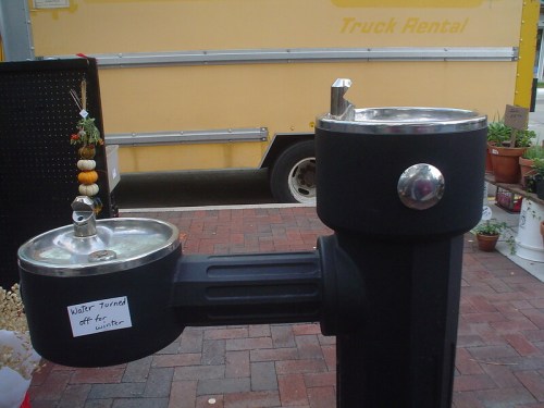 A water fountain with a sign reading "Water turned off for winter," next to a yellow truck and potted plants.