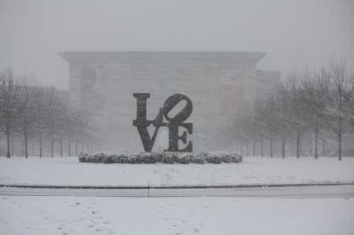 A large "LOVE" sculpture stands in a snowy landscape, surrounded by bare trees and a blurred building in the background.