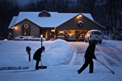 Two people shoveling snow in front of a warmly lit house decorated for the holidays, with a snowy driveway.