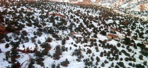 Aerial view of a snowy landscape with scattered trees and a small building amidst red soil and rocky terrain.