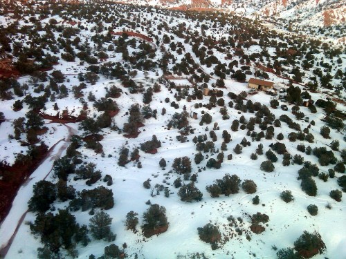 Aerial view of a snowy landscape with scattered trees and a few buildings in a mountainous area.