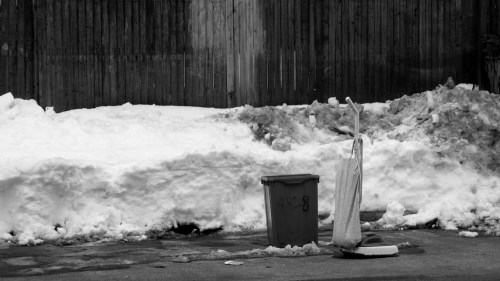 A trash can and a snow shovel stand against a backdrop of snow and a wooden fence in a black-and-white scene.
