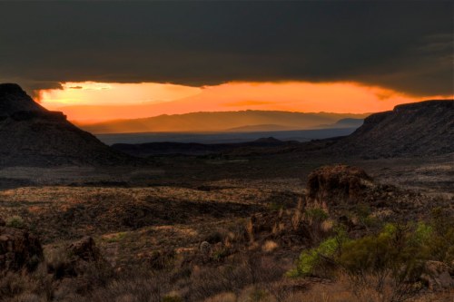A dramatic sunset over a rugged desert landscape, with dark clouds and distant mountains silhouetted against the orange sky.