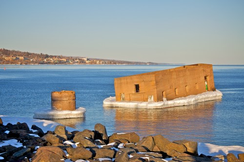A partially submerged concrete structure on icy water, with a rocky shoreline and a distant town in the background.