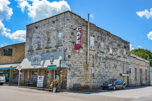 Historic stone building with a café sign, featuring shops and a blue sky with fluffy clouds.