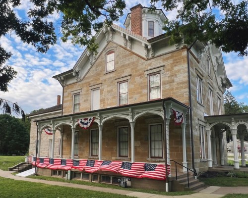 Historic stone house adorned with American flags and bunting, set against a blue sky with scattered clouds.