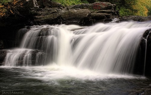 A serene waterfall cascades over rocky ledges, surrounded by lush greenery and autumn leaves.