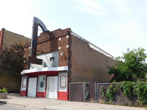 Abandoned theater building with a vintage marquee, red and white facade, and overgrown greenery nearby.