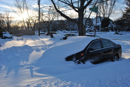 A car almost completely buried in snow, with a sunset in the background and snow-covered trees lining the street.
