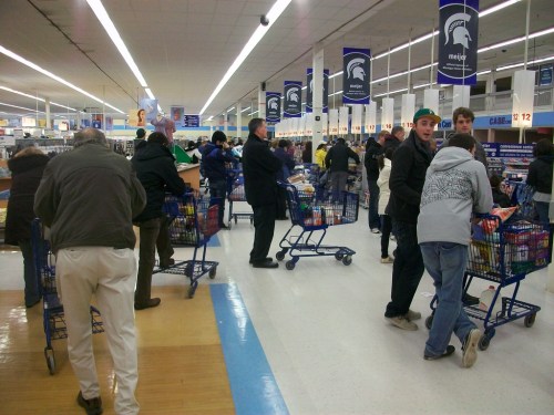 A busy grocery store aisle with shoppers pushing carts and browsing products under bright lights.