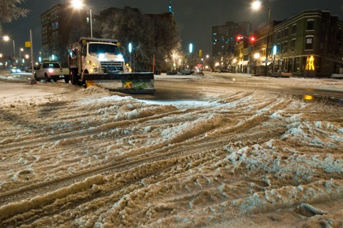 A snowplow clears a snowy street at night, with city buildings and streetlights in the background.