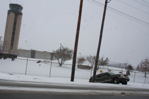 A green SUV is partially in a snow-covered ditch near a fenced area and a tall airport control tower in the background.
