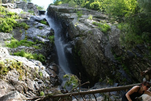 A waterfall cascading down rocky cliffs, surrounded by lush greenery and a person exploring the area nearby.