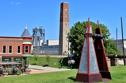 A park scene featuring a large sculpture, historic buildings, and a tall brick tower against a clear blue sky.