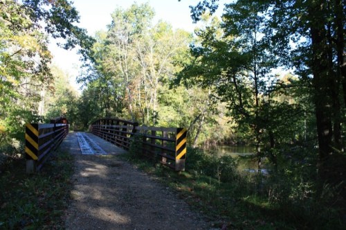 A wooden bridge over a calm river, surrounded by lush green trees and a clear blue sky.