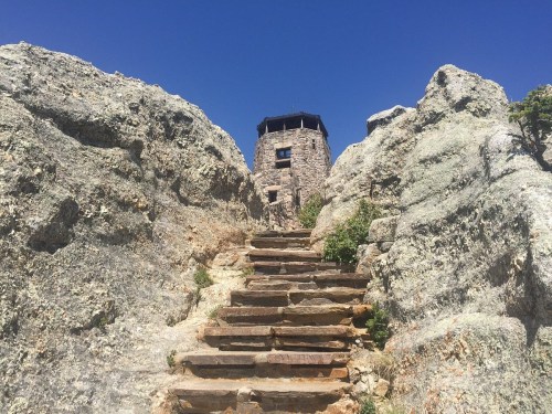 Stone steps lead up to a tower nestled between large rocky formations under a clear blue sky.