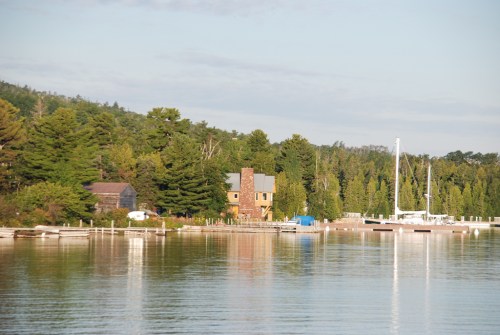A serene lakeside view featuring a house, dock, and sailboats surrounded by lush trees and calm water.