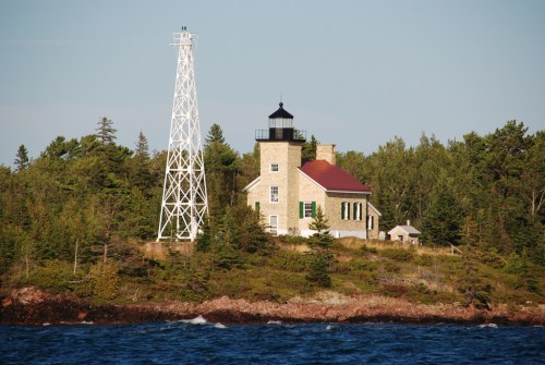 A lighthouse with a red roof stands on a rocky shore, surrounded by trees and water under a clear blue sky.