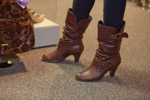 A close-up of a person's legs wearing brown slouchy heeled boots, standing on a carpeted floor near a handbag.