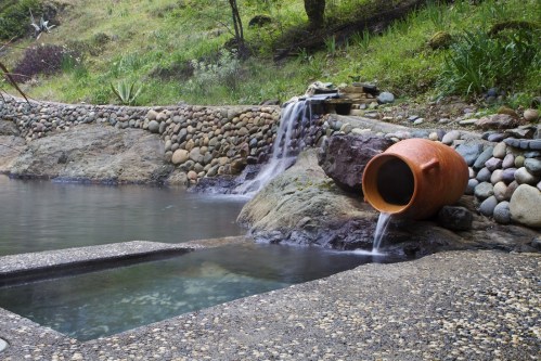 A serene natural hot spring with a stone border, a clay pot, and gentle waterfalls surrounded by greenery.