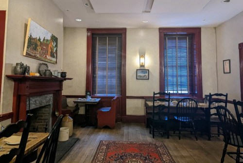 Cozy restaurant interior with wooden floors, red accents, and a fireplace, featuring tables and chairs by the windows.