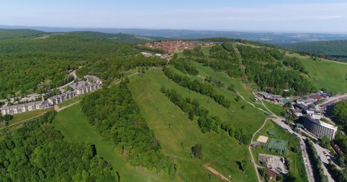 Aerial view of a lush green hillside with buildings and a distant town surrounded by trees and rolling hills.