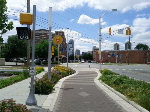 A city street view with traffic signals, a pedestrian path, and buildings in the background under a partly cloudy sky.