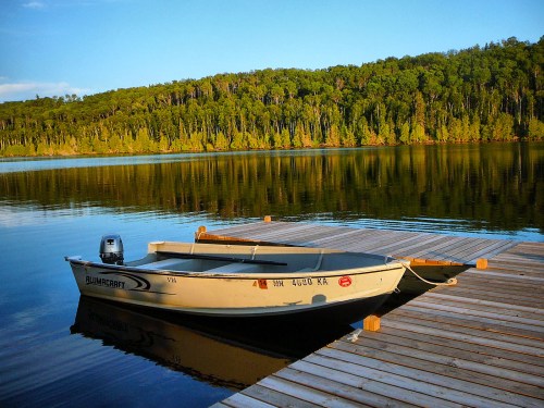 A small boat is docked on a calm lake, surrounded by lush green trees and a clear blue sky.