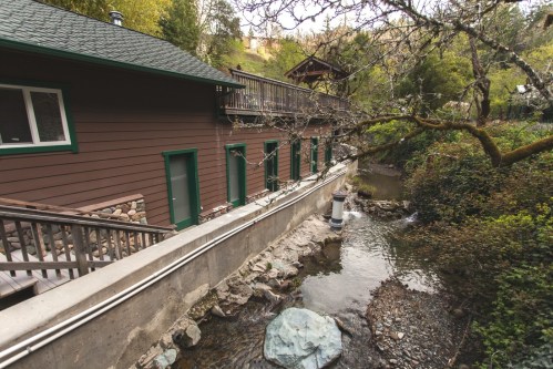 A rustic building beside a small stream, surrounded by greenery and trees, with a wooden deck above.